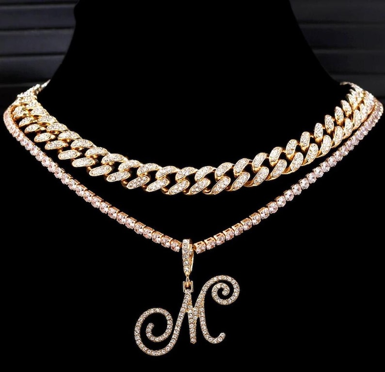 Peut inclure: Ensemble de colliers dor&eacute;s &agrave; trois niveaux. La couche sup&eacute;rieure est une cha&icirc;ne &eacute;paisse orn&eacute;e de diamants. La couche interm&eacute;diaire est un d&eacute;licat collier de tennis. La couche inf&eacute;rieure pr&eacute;sente un pendentif avec la lettre "M" incrust&eacute;e de diamants. Le fond est noir.
