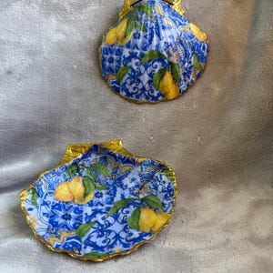 Może przedstawiać: Dwie ozdobne muszle z niebiesko-białym wzorem płytek i złotymi akcentami. Muszle mają motyw cytrynowy i idealnie nadają się do dodania odrobiny śródziemnomorskiego uroku do wystroju domu.