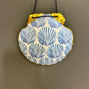 May include: A white seashell with a blue and white pattern, accented with gold trim, hangs from a black cord.