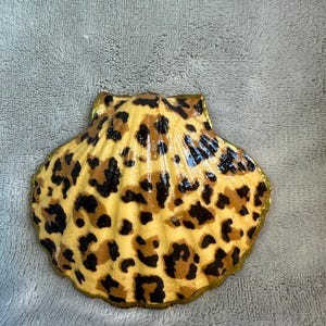 Könnte beinhalten: Eine dekorative Muschel, bemalt mit einem Leopardenmuster. Die Muschel ist gelb mit braunen und schwarzen Flecken und hat einen goldfarbenen Rand. Die Muschel liegt auf einer grauen Oberfläche.