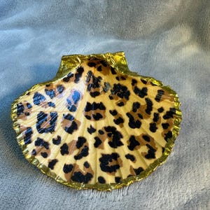 May include: A decorative seashell with a leopard print pattern. The shell has a tan base with brown and black spots. The edges are trimmed with gold leaf, adding a touch of elegance to this unique piece.