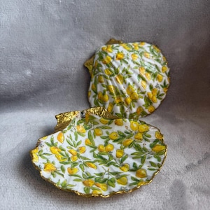 May include: Two white ceramic seashells with a gold rim. The shells are decorated with a lemon print.