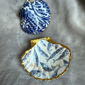 May include: Two decorative seashell dishes. One features a blue and white floral pattern, the other has a white background with blue fish illustrations. Both shells have gold-painted edges, adding a touch of elegance.