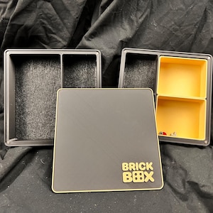 Könnte beinhalten: Eine schwarze Aufbewahrungsbox mit goldfarbenem Rand und den Worten "BRICK BOX" in Gold. Die Box ist geöffnet und zeigt zwei Fächer. Eine weitere Box mit zwei gelben Fächern ist ebenfalls sichtbar. Die Boxen scheinen für die Organisation kleiner Gegenstände konzipiert zu sein.