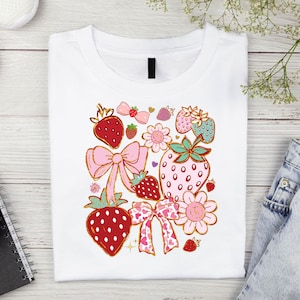 以下が含まれることがあります： ピンク、赤、緑のイチゴのデザインが施された白いTシャツ。イチゴはピンクのリボンと花で囲まれています。