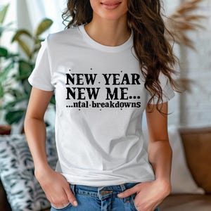 May include: A white short-sleeved t-shirt featuring black text that reads NEW YEAR NEW ME... ...ntal breakdowns, accented with small star symbols. The shirt has a crew neckline and is paired with blue denim jeans, creating a casual look.
