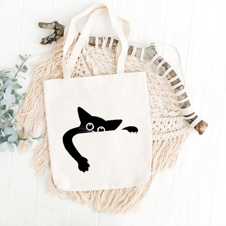 May include: A cream-colored tote bag with a black cat design. The cat's head and paw peek over the top, with large eyes and a long, curved tail. The bag has two cream-colored handles.