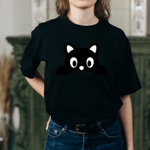May include: Black t-shirt featuring a cartoon cat design. The cat is black with white accents on the ears and a white nose. The cat's eyes are large and round. The shirt is made of a soft material and has short sleeves.