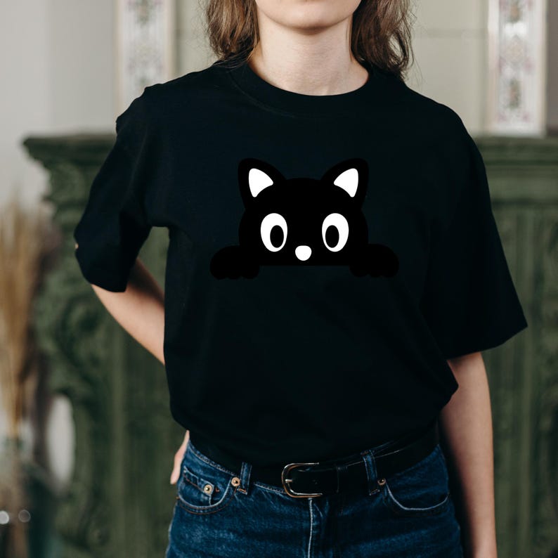 May include: Black t-shirt featuring a cartoon cat design. The cat is black with white accents on the ears and a white nose. The cat's eyes are large and round. The shirt is made of a soft material and has short sleeves.