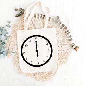 May include: A white canvas tote bag with a black clock face design. The clock face shows the time as 12:00.
