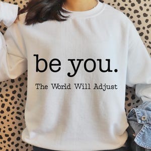 Puede incluir: Sudadera blanca con el texto "be you." sobre "The World Will Adjust" en negro, con letras sans-serif. La sudadera tiene cuello redondo y mangas largas.