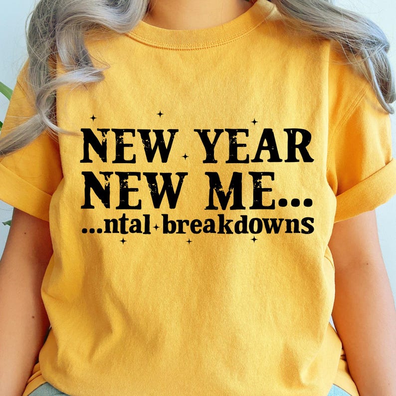 May include: A mustard yellow short-sleeved t-shirt featuring black text that reads NEW YEAR NEW ME... ...ntal breakdowns, accented with small four-pointed star symbols scattered throughout the lettering on the front of the garment.