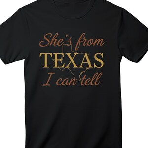 May include: Black t-shirt featuring the text "She's from TEXAS I can tell" in a brown script font. The word "TEXAS" is in a gold block font with a gold outline of the state of Texas.
