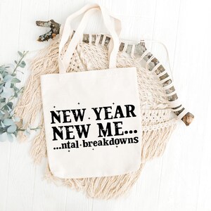 May include: A cream-coloured tote bag featuring the text "NEW YEAR NEW ME... ...ntal breakdowns" in black. The bag has two handles and is set against a neutral-toned macrame backdrop.