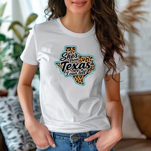 May include: White t-shirt with a graphic of the state of Texas and the text "She's from Texas I can tell". The Texas outline is filled with a leopard print pattern, outlined in teal, and the text is in black.