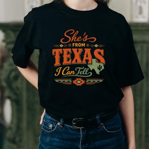 May include: Black t-shirt featuring the text "She's from Texas I Can Tell" in orange and white. The design includes a Texas outline and a cactus. The shirt is worn with blue jeans.