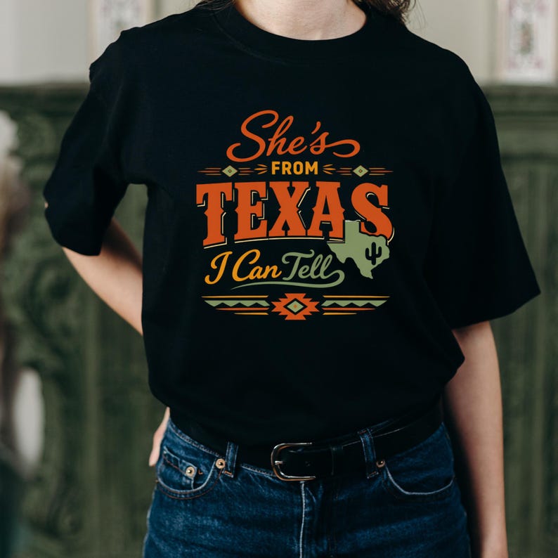 May include: Black t-shirt featuring the text "She's from Texas I Can Tell" in orange and white. The design includes a Texas outline and a cactus. The shirt is worn with blue jeans.