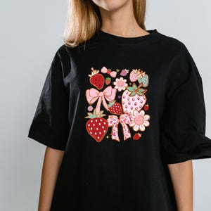 以下が含まれることがあります： イチゴ、リボン、花をカラフルなグラフィックプリントした黒いTシャツです。