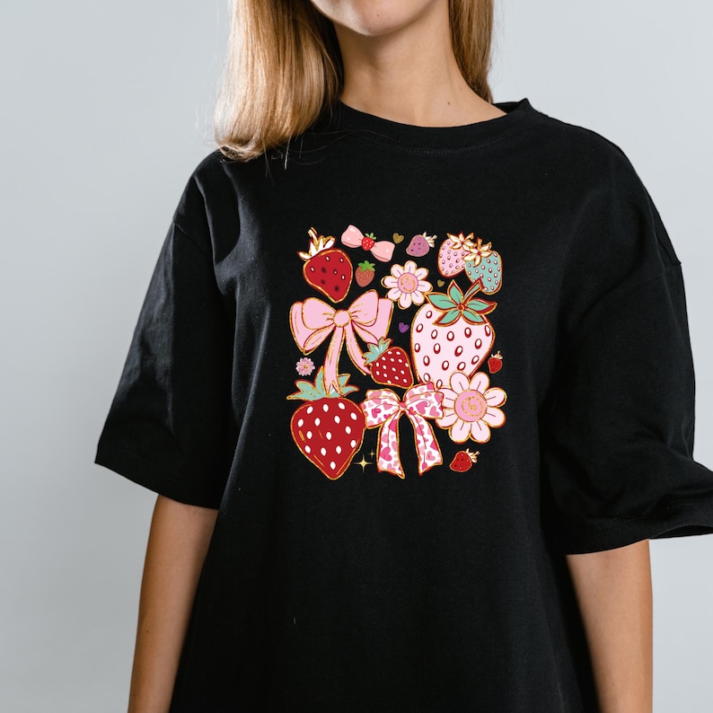 以下が含まれることがあります： イチゴ、リボン、花をカラフルなグラフィックプリントした黒いTシャツです。