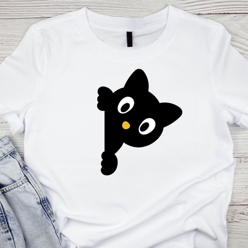 May include: White t-shirt featuring a black cat graphic peeking from the side. The cat has large white eyes, a small yellow nose, and is designed in a cartoon style. The shirt is made of a soft material.