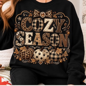 Puede incluir: Una sudadera negra de manga larga con un estampado gráfico que incluye el texto COZY SEASON. El diseño presenta un lazo con estampado de leopardo, tres calabazas con patrones de leopardo, lunares y cuadros, y detalles dorados.