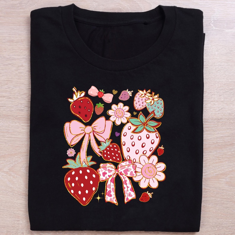 以下が含まれることがあります： イチゴ、リボン、花をモチーフにしたカラフルなグラフィックプリントが施された黒いTシャツ。デザインにはピンク、赤、黄、緑の色が使われています。