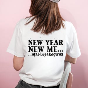 May include: White t-shirt with the text "NEW YEAR NEW ME... ...ntal breakdowns" in black. The shirt is short-sleeved and has a crew neck. The image shows the back of the shirt.