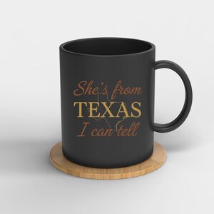 May include: A black ceramic mug with the text "She's from TEXAS I can tell" in brown and gold. The mug is on a round, light brown coaster. The mug is a standard size.