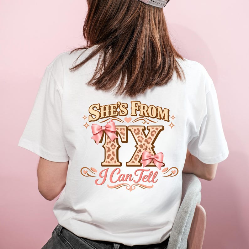 May include: White t-shirt featuring the text "She's From TX I Can Tell". The letters "TX" are in a leopard print design, embellished with pink bows. The shirt is a casual style, suitable for everyday wear.