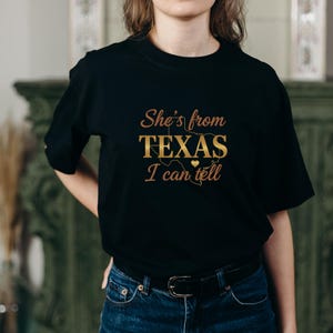 May include: Black t-shirt featuring the gold text "She's from TEXAS I can tell", with a heart and a map of Texas. The shirt has a crew neck design.