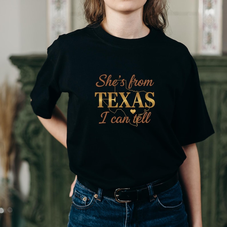 May include: Black t-shirt featuring the gold text "She's from TEXAS I can tell", with a heart and a map of Texas. The shirt has a crew neck design.