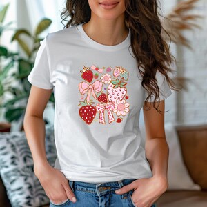 以下が含まれることがあります： 白いTシャツに、イチゴ、リボン、花をモチーフにしたカラフルなグラフィックデザインが施されています。デザインはTシャツの中央に配置されています。
