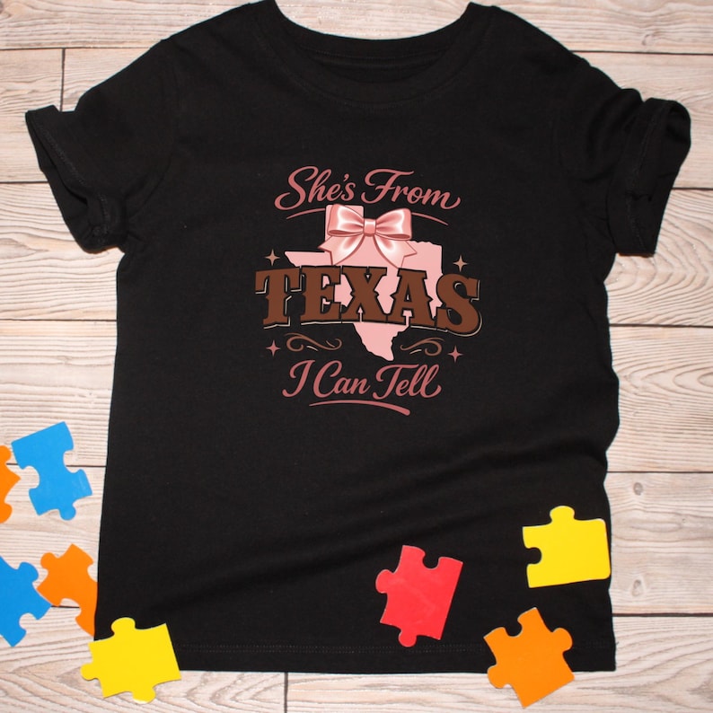 May include: Black t-shirt featuring the text "She's From Texas I Can Tell" in pink and brown. The design includes a pink outline of Texas and a bow. Colourful puzzle pieces are scattered around the shirt.