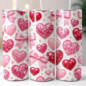 May include: Three stainless steel tumblers with a Valentine's Day theme. The tumblers are covered in a pattern of pink hearts, bows, and glitter on a white background. Each tumbler has a straw.