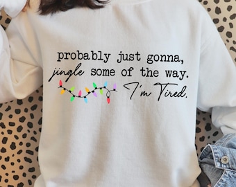 May include: White sweatshirt with the text "probably just gonna, jingle some of the way. I'm Tired." A string of colorful Christmas lights is below the text. The sweatshirt is a crew neck style.