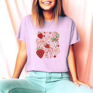 以下が含まれることがあります： 薄い紫色のTシャツに、イチゴ、リボン、花がピンク、赤、緑、金でデザインされています。