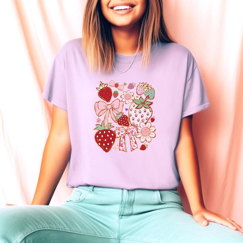 以下が含まれることがあります： 薄い紫色のTシャツに、イチゴ、リボン、花がピンク、赤、緑、金でデザインされています。