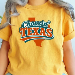 May include: Mustard yellow t-shirt featuring the text "Choosin' TEXAS" in a teal, orange, and white design. The design includes a stylized outline of the state of Texas. The shirt has rolled-up sleeves.
