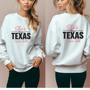 May include: White sweatshirt featuring the text "She's From Texas I Can Tell" in black and pink. The image shows the front and back of the sweatshirt. The sweatshirt is a casual style.