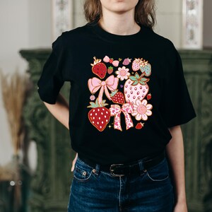 以下が含まれることがあります： イチゴ、リボン、花をモチーフにしたカラフルなグラフィックプリントが施された黒いTシャツ。デザインにはピンク、赤、緑、黄色の色が使われています。