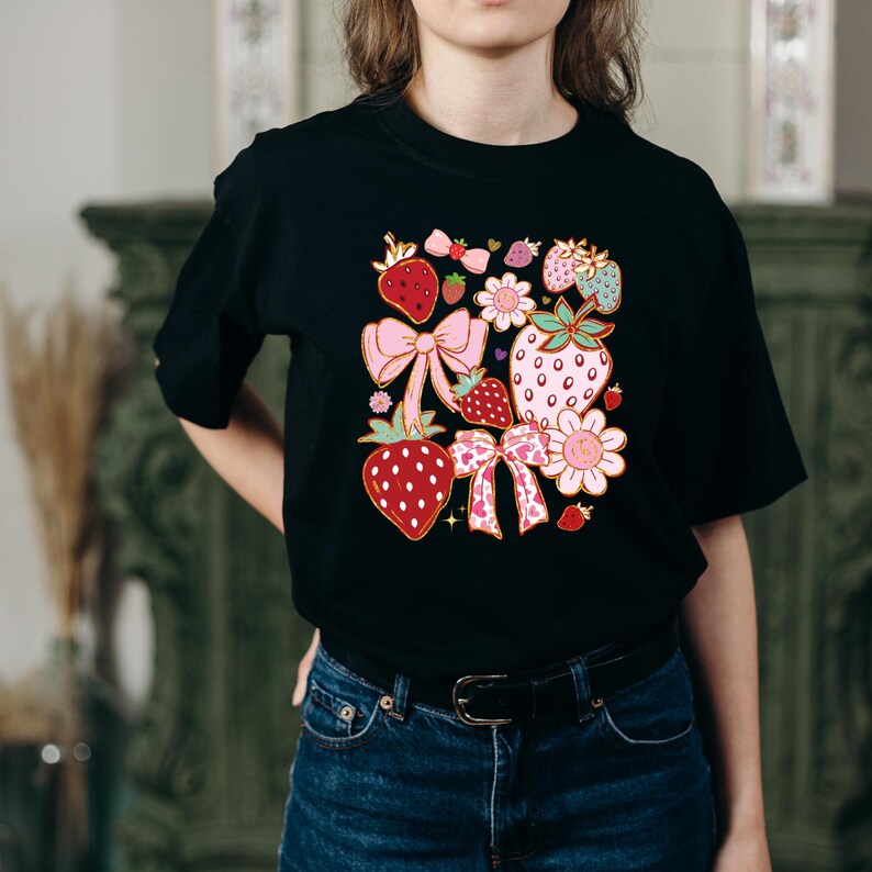 以下が含まれることがあります： イチゴ、リボン、花をモチーフにしたカラフルなグラフィックプリントが施された黒いTシャツ。デザインにはピンク、赤、緑、黄色の色が使われています。
