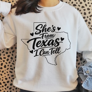 May include: White sweatshirt featuring a black outline of the state of Texas with the words "She's From Texas I Can Tell". The design includes hearts and a rectangular border around "She's". The sweatshirt is a casual style.