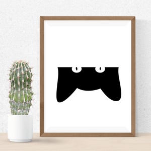 May include: A framed art print featuring a minimalist black cat design. The cat's silhouette is black with two white oval eyes, set against a white background. The frame is a warm brown color, and a cactus in a white pot is visible on the left.