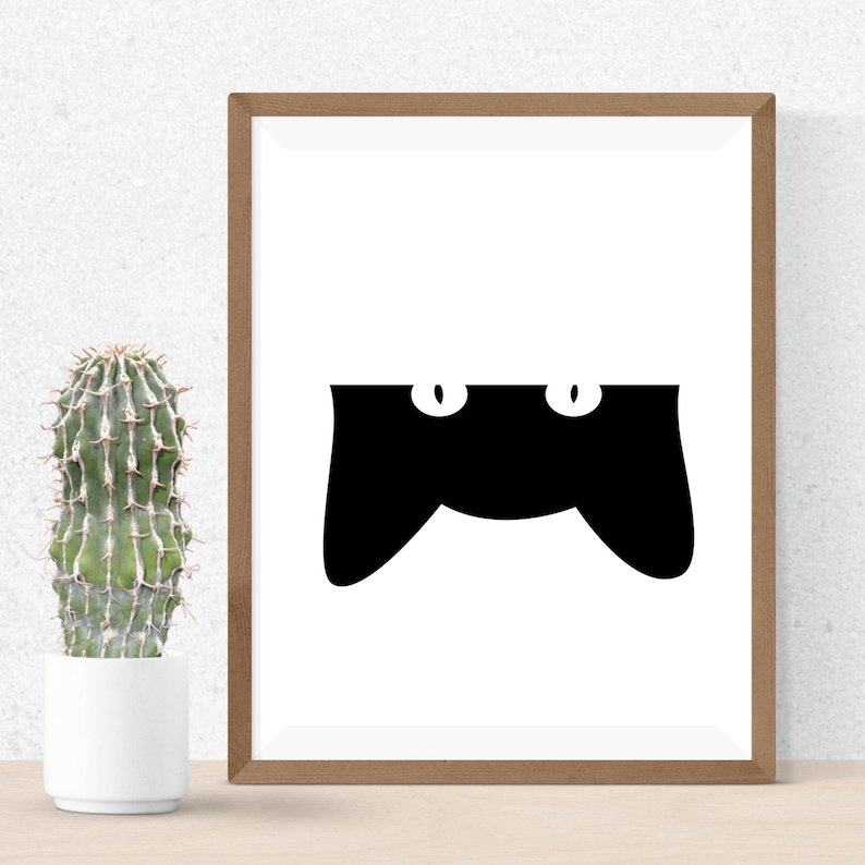 May include: A framed art print featuring a minimalist black cat design. The cat's silhouette is black with two white oval eyes, set against a white background. The frame is a warm brown color, and a cactus in a white pot is visible on the left.