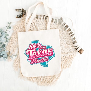 May include: Cream tote bag featuring the text "She's from Texas I Can Tell" in pink and teal. The text is in a stylized font and set against a teal outline of the state of Texas. The bag has cream-colored handles and is made of canvas.
