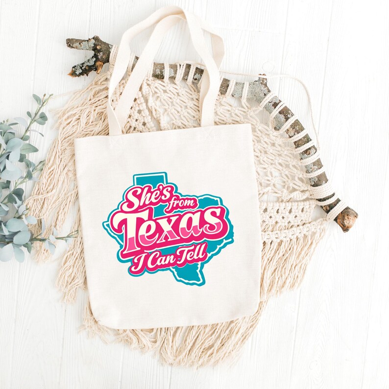 May include: Cream tote bag featuring the text "She's from Texas I Can Tell" in pink and teal. The text is in a stylized font and set against a teal outline of the state of Texas. The bag has cream-colored handles and is made of canvas.