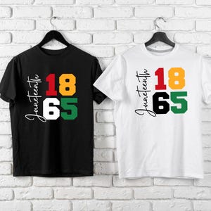 May include: Two t-shirts, one black and one white, each with the word "Juneteenth" in cursive and the numbers "18" and "65" in red, yellow, black, and green. The shirts are hanging on black hangers.