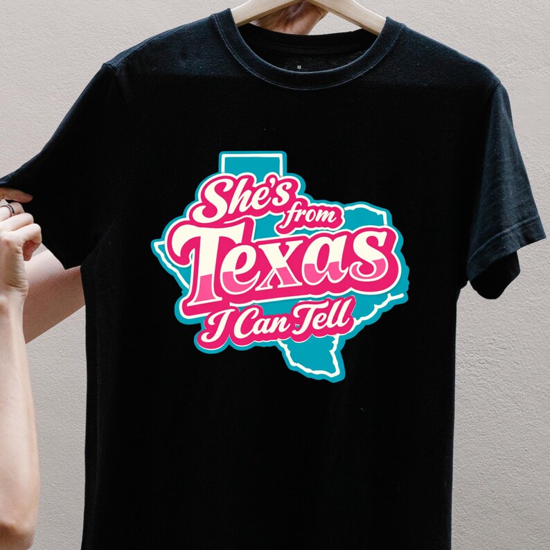 May include: Black t-shirt with a graphic of the state of Texas in turquoise, with the text "She's from Texas I Can Tell" in pink and white. The design has a retro style.