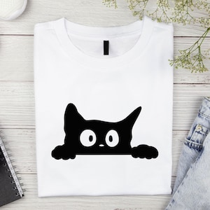 May include: White t-shirt featuring a black cat graphic. The cat's head and paws peek over the bottom edge, with large white eyes and a small nose. The shirt is folded neatly on a light wood-grain surface.