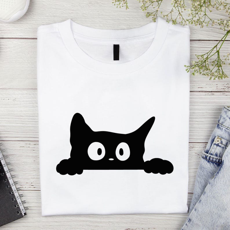 May include: White t-shirt featuring a black cat graphic. The cat's head and paws peek over the bottom edge, with large white eyes and a small nose. The shirt is folded neatly on a light wood-grain surface.